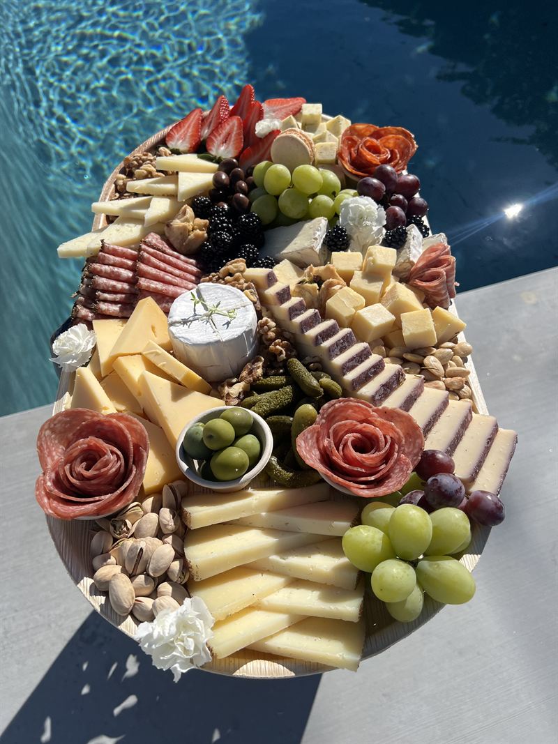 Large charcuterie board loaded with cheeses, meats and grapes beside a pool